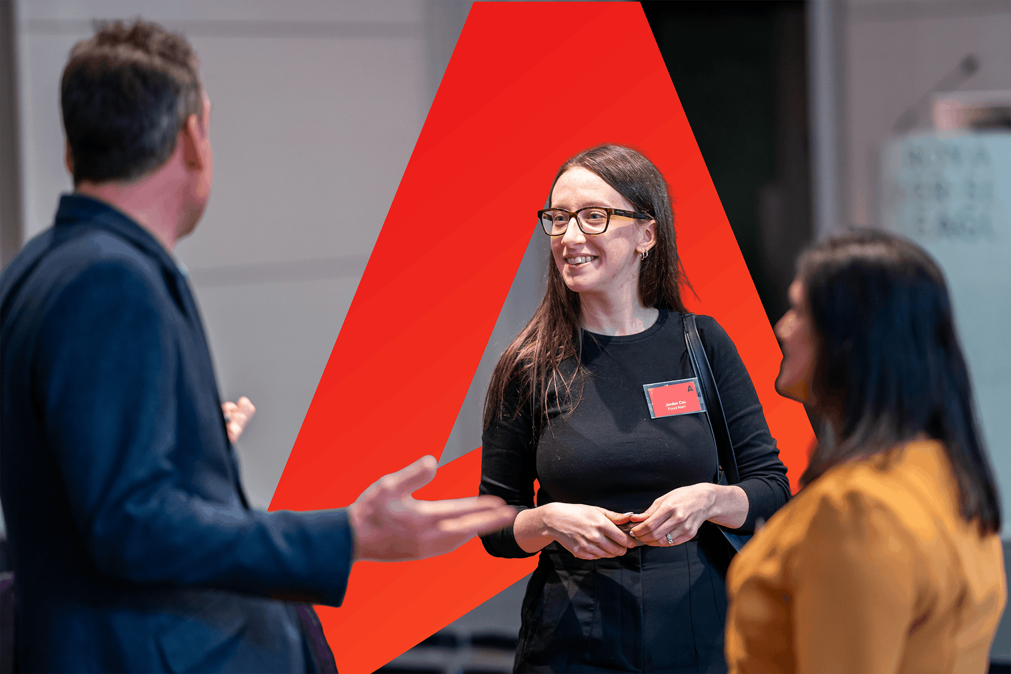 Business professionals engaged in conversation at a networking event, featuring a woman smiling and wearing glasses, a name tag, and two colleagues discussing in the background. A prominent red geometric shape adds visual interest to the image.