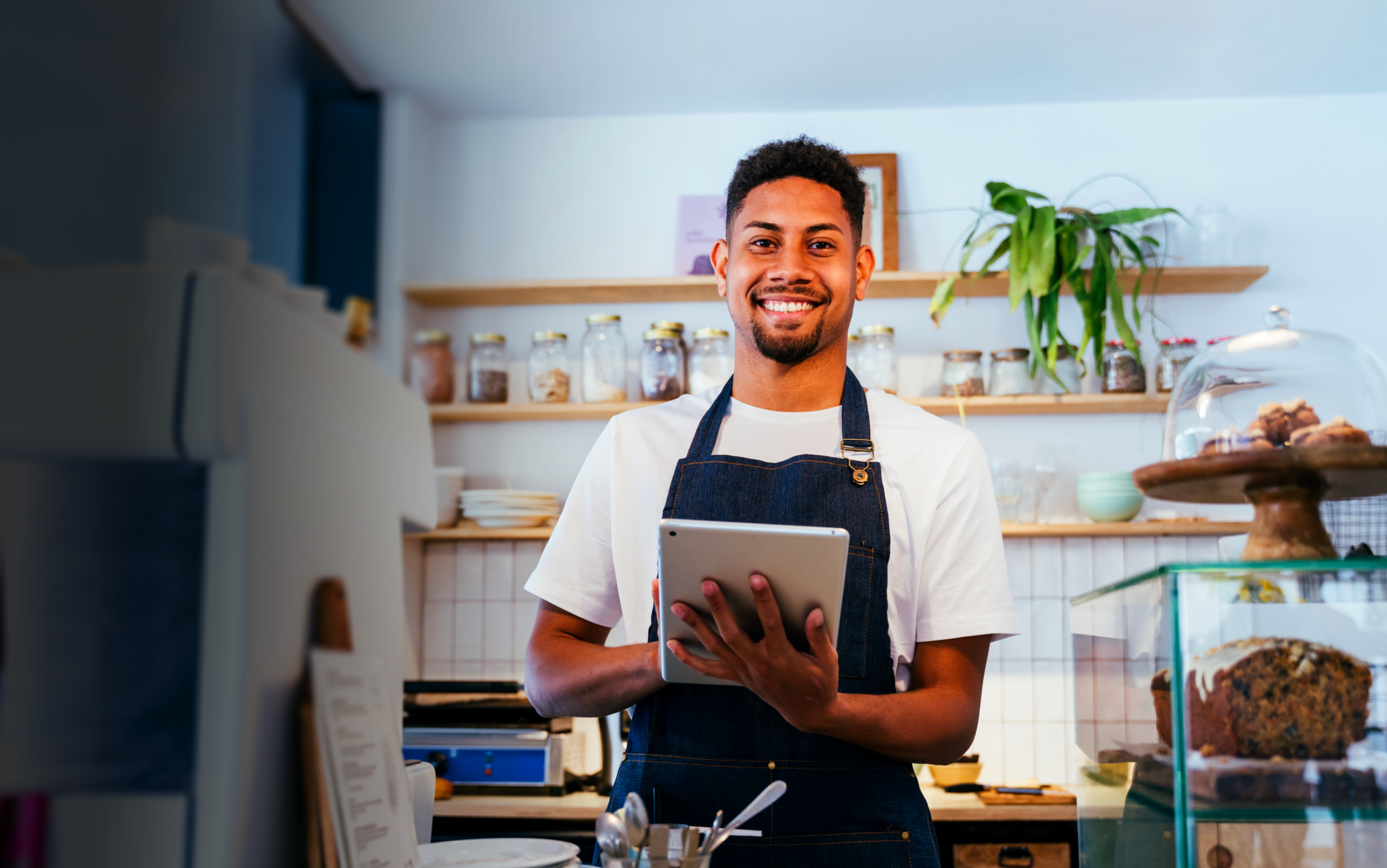 Smiling barista in a cozy café holding a tablet, surrounded by jars of ingredients and baked goods, wearing a denim apron and a casual t-shirt, showcasing a welcoming atmosphere for customers.