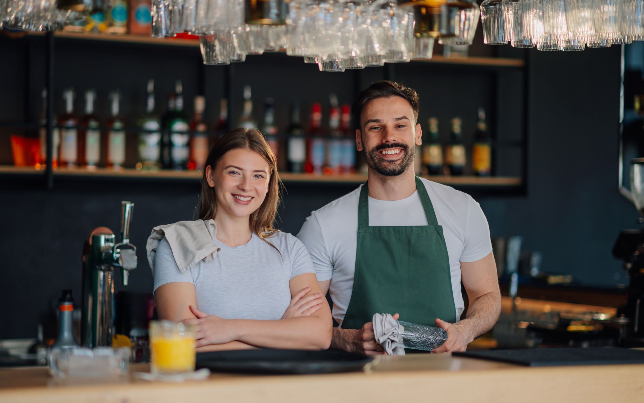 Smiling bar staff in a modern bar setting, with a diverse selection of liquor bottles in the background, showcasing a welcoming atmosphere for patrons.